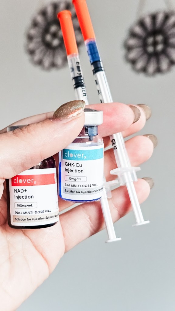 Hand holding CloveRX NAD+ and GHK-Cu injection vials prescribed through EllieMD telehealth with two insulin syringes for subcutaneous peptide injections